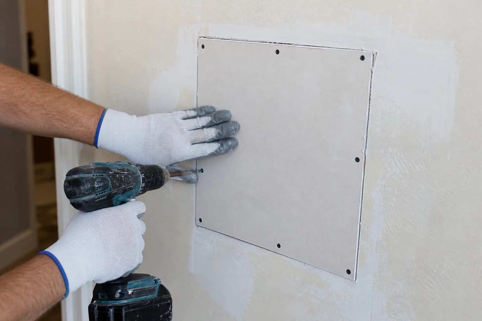 Contractor's gloved hands fastening a new drywall patch into a wall opening with screws along the edges, ready for taping, during a water-damage drywall repair