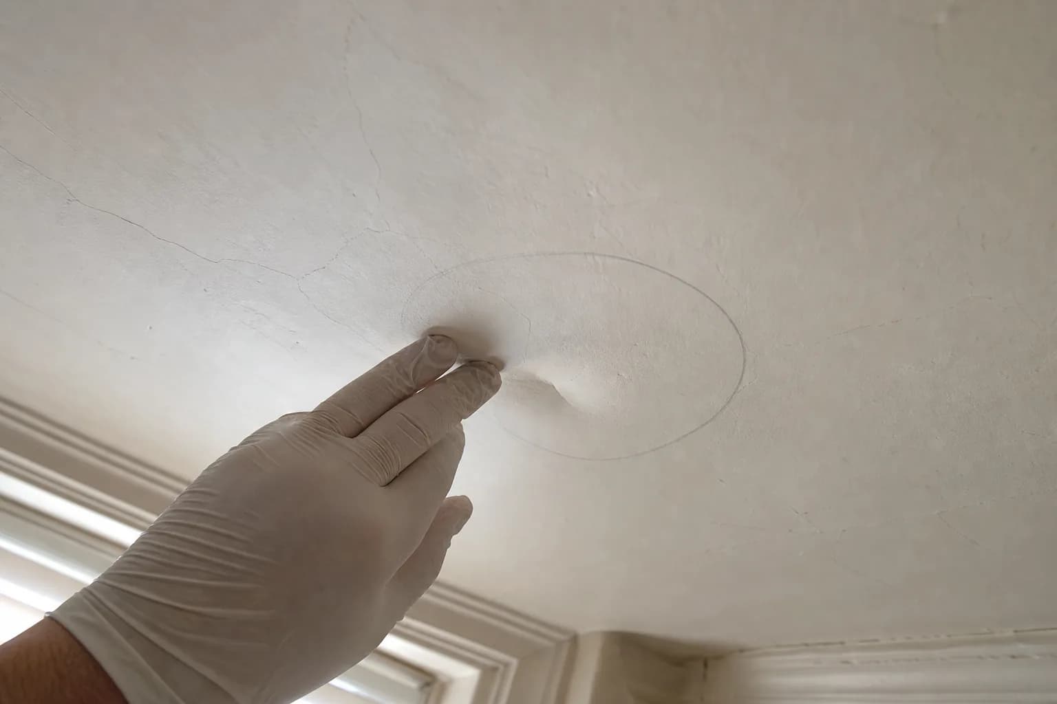 A gloved hand gently pressing a pencil-circled bulge on an older plaster ceiling near a network of hairline cracks, a simple check for loose plaster before repair